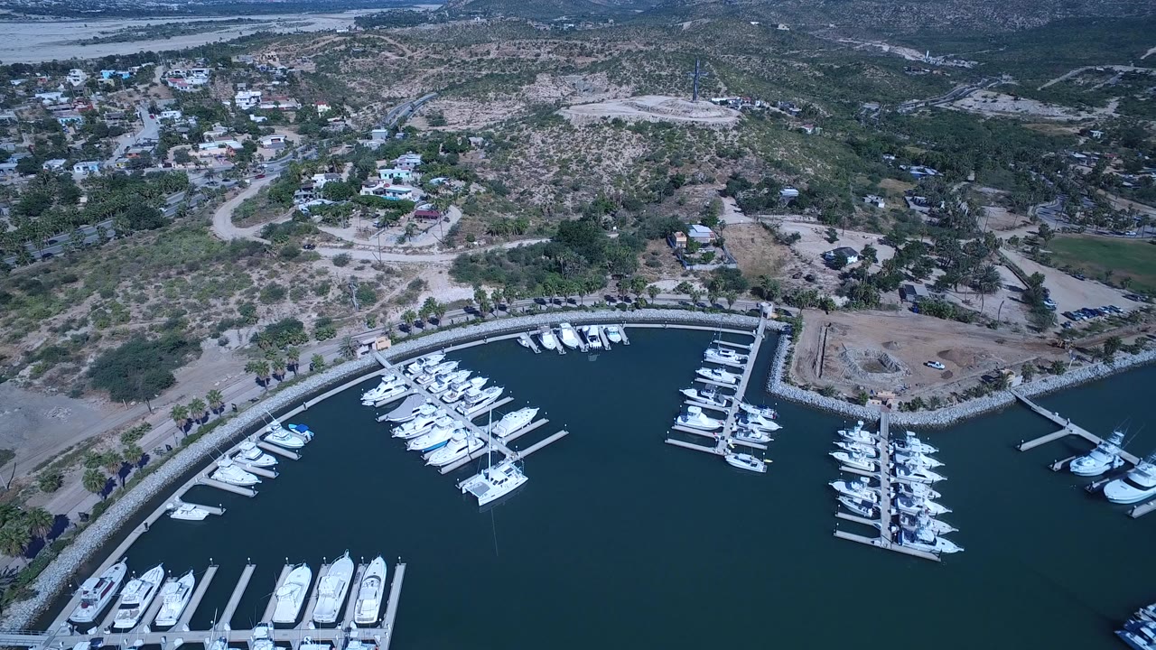 San Jose Del Cabo aerial frame showing a marina packed with white boats, docks along a curved wall, hillside homes, roads, trees, and no clearly visible people.