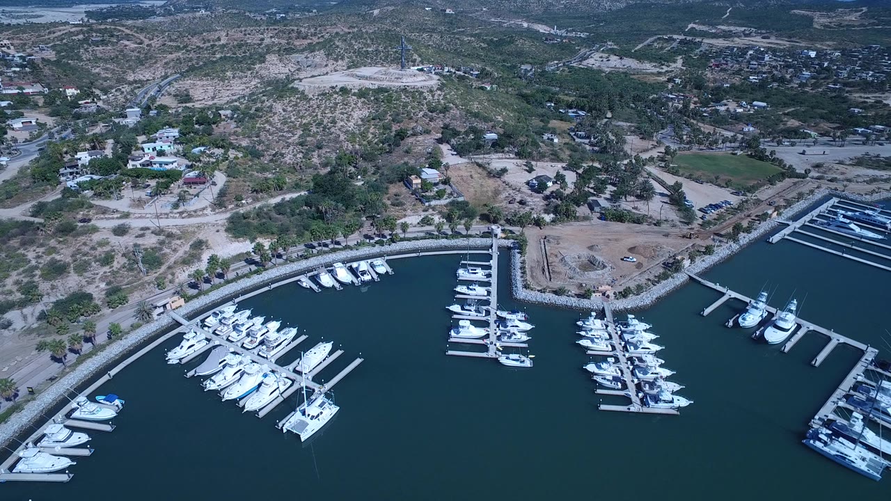 High aerial view in San Jose Del Cabo showing white boats moored around marina fingers, a breakwater edge, hillside roads, and no visible people.