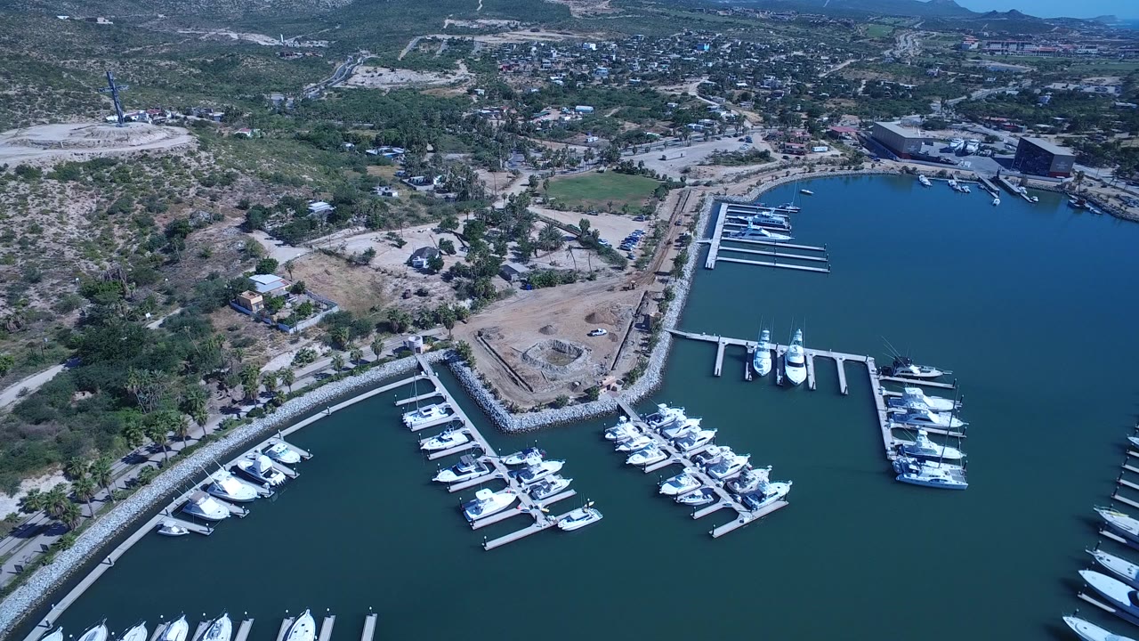 High aerial frame over a San Jose del Cabo marina, with white yachts in foreground slips, a curved breakwater at left, dry shoreline lots inland, and no visible people.