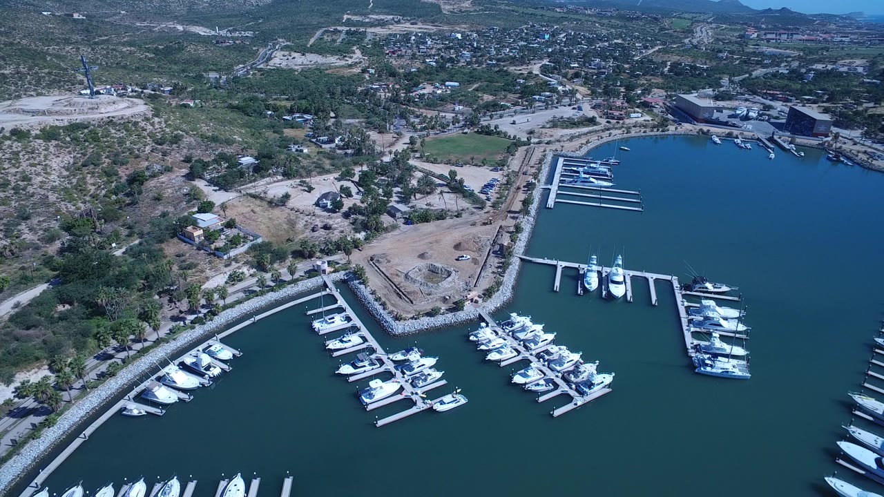 Aerial view in San Jose del Cabo showing marina docks grouped around teal water, a dry lot on the shore, scattered inland buildings, and no visible people.