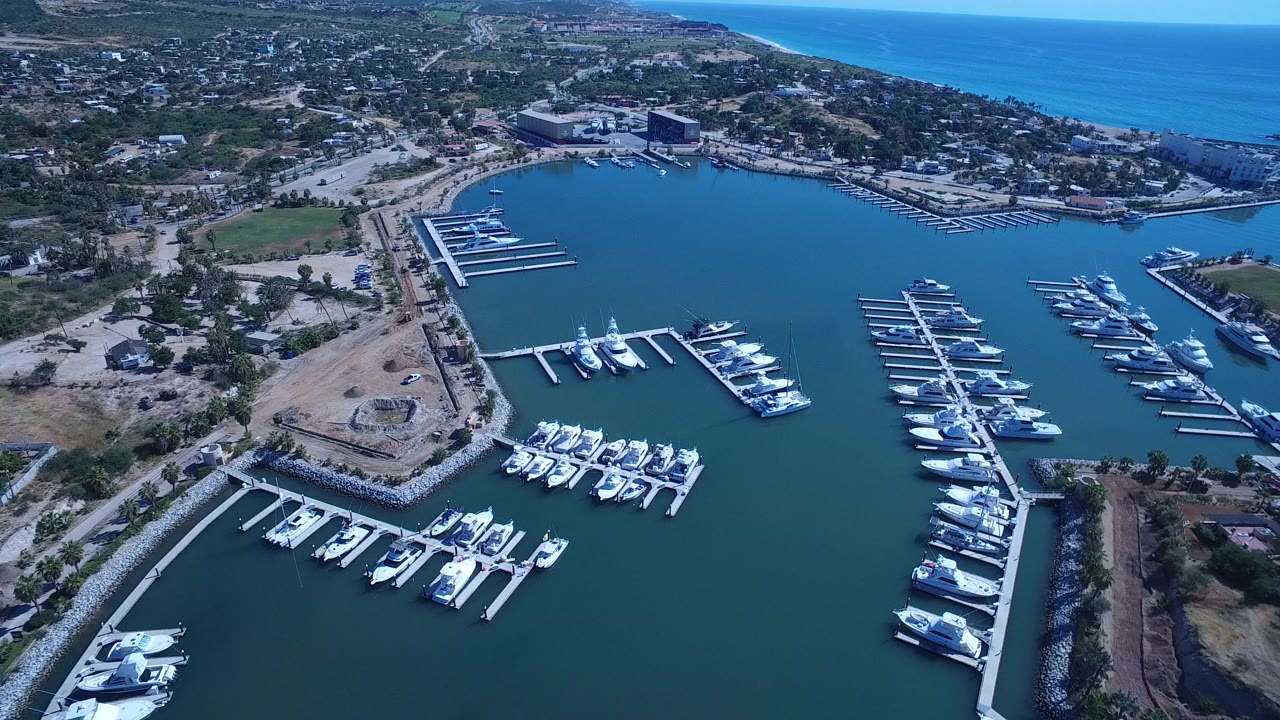 Opening aerial frame over San Jose del Cabo, Mexico, showing white yachts moored in a teal marina bay, dry shoreline work areas, low buildings, roads, and the ocean horizon with no visible people.