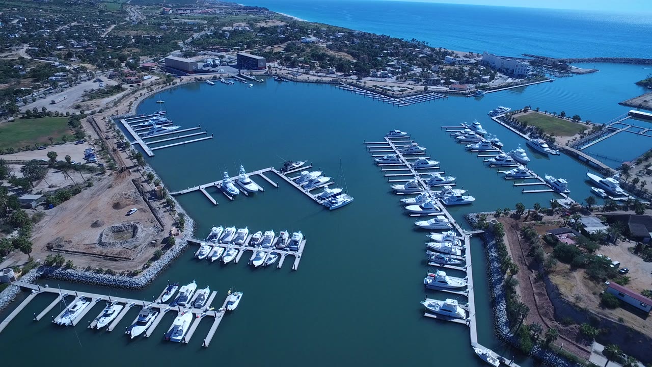 Aerial frame over San Jose del Cabo showing white boats on marina docks, turquoise bay water, shoreline roads, coastal buildings, open ocean, and no visible people.