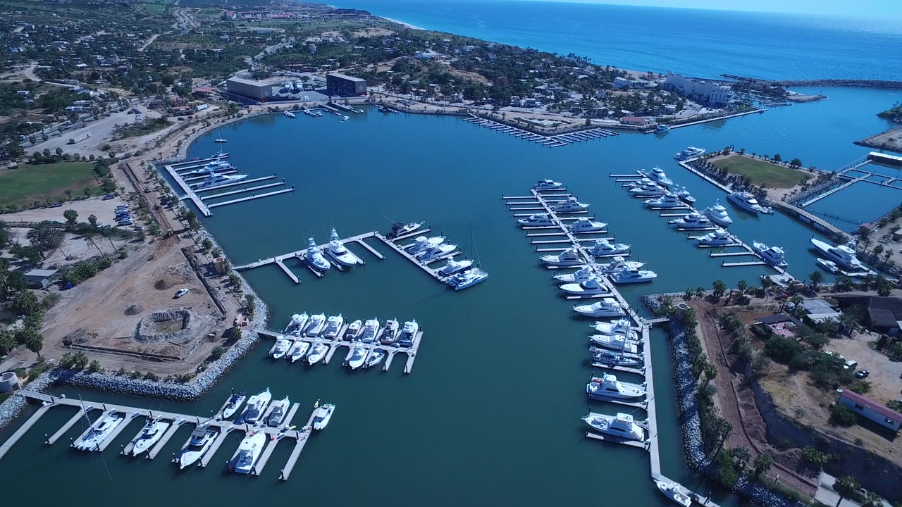 High aerial view in San Jose del Cabo with yacht docks spread across the bay, a rocky edge in the foreground, roads and buildings inland, ocean at the horizon, and no visible people.
