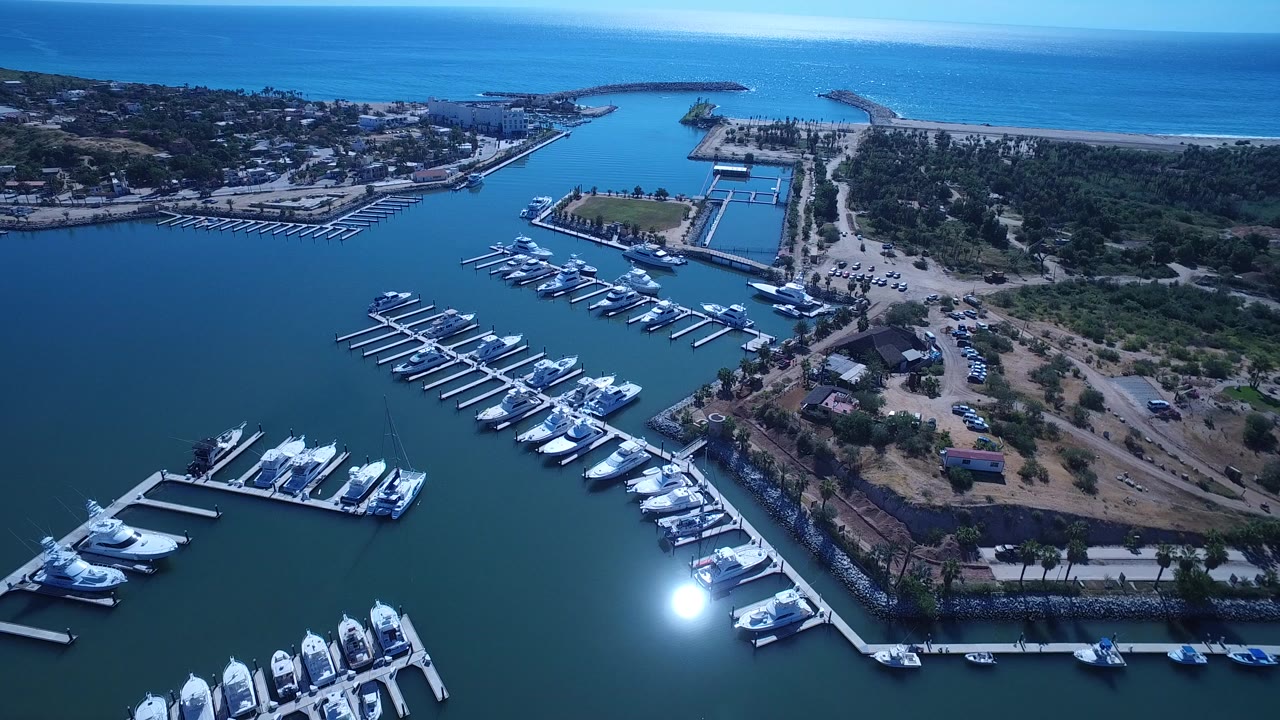 Opening aerial frame over a San Jose del Cabo marina with white boats at docks, shoreline roads, a beach and blue sea beyond, and no visible people.