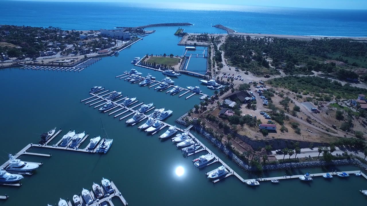Drone-like aerial over marina slips, boats, breakwaters, beach, and sea.