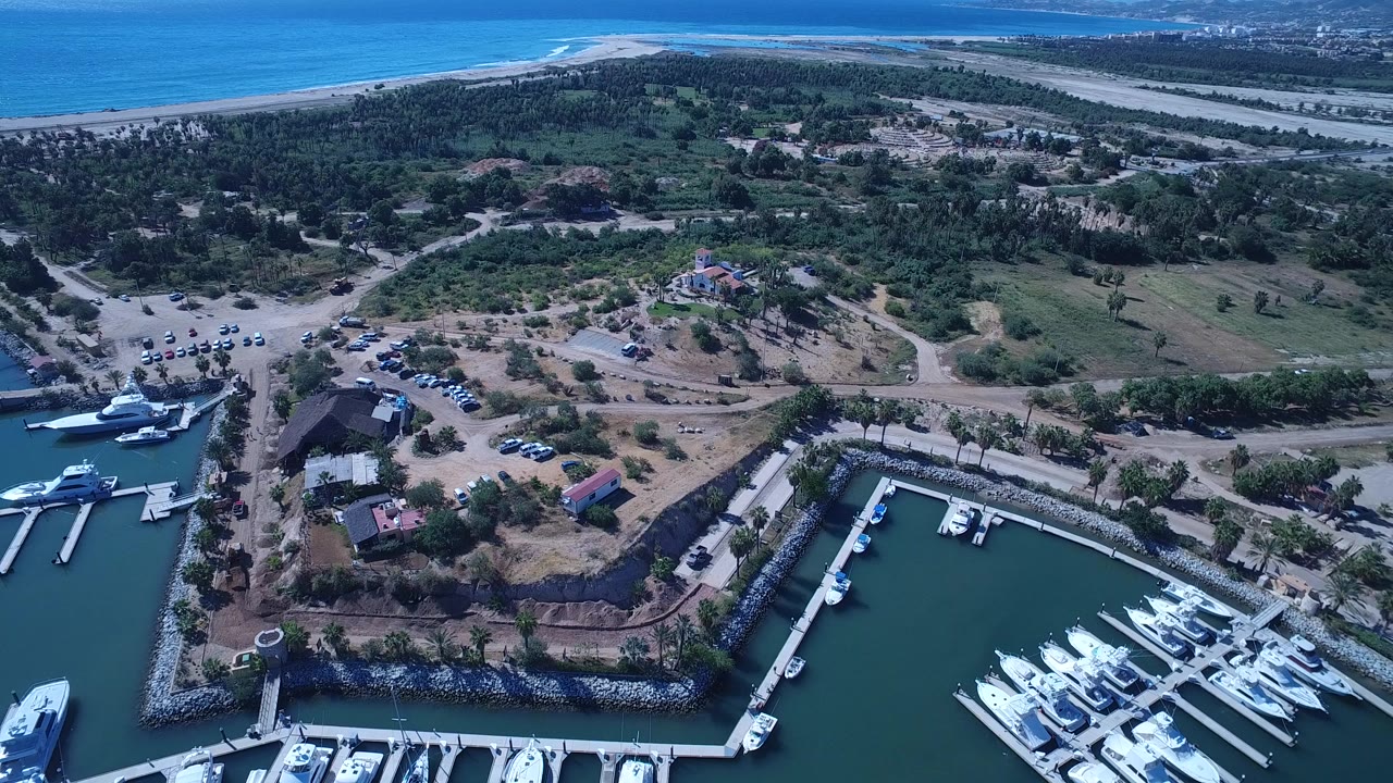 Opening aerial frame over San Jose del Cabo shows a marina basin with white boats at the lower edge, roads and trees through the center, ocean shoreline in back, and no visible people.