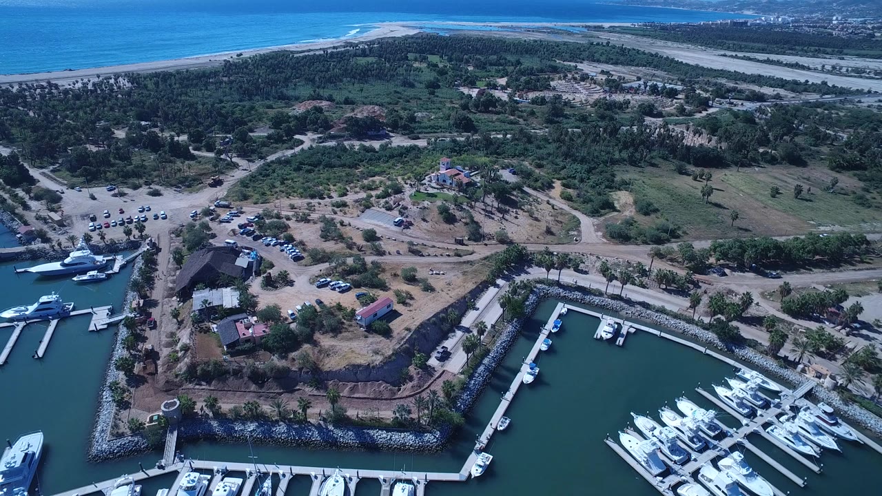 San Jose del Cabo aerial frame shows more of the marina entry at left, docked boats at lower right, a road grid through dry greenery, and the ocean shoreline behind, with no visible people.
