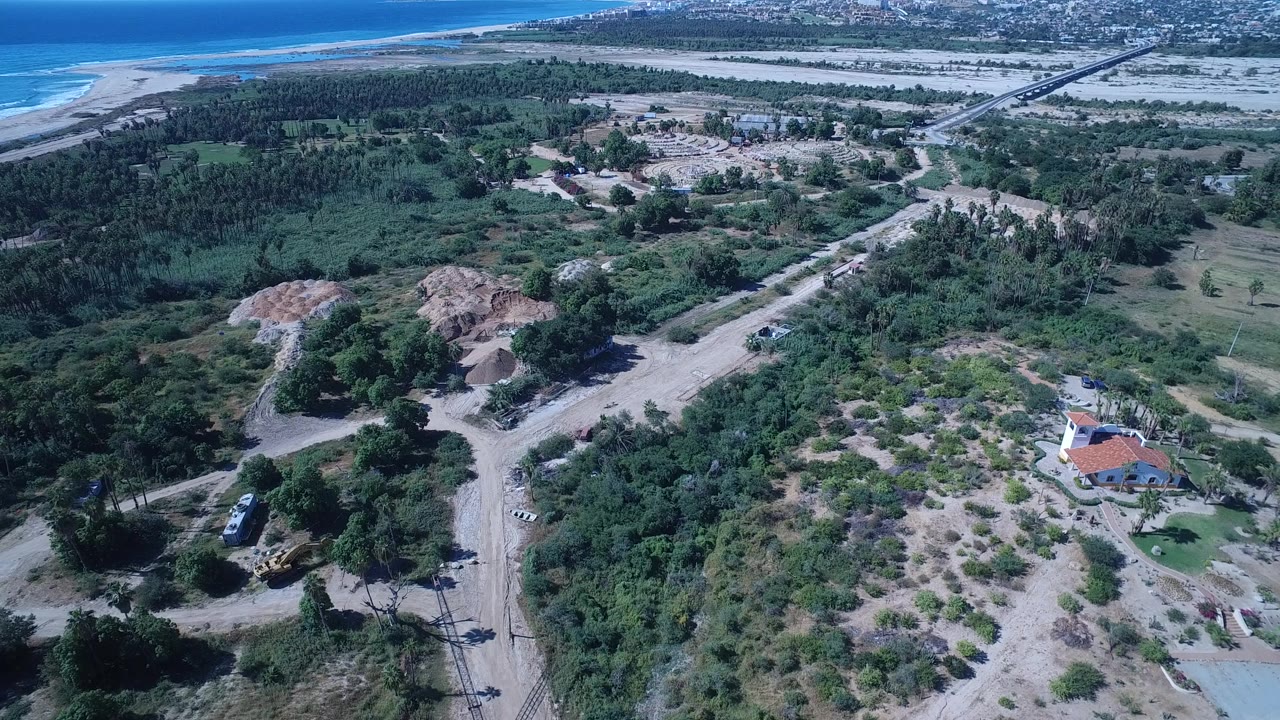 Second aerial frame over San Jose del Cabo, Mexico, with the red-roofed building lower right, sandy tracks through scrub, dune mounds midframe, ocean beyond, and no visible people.