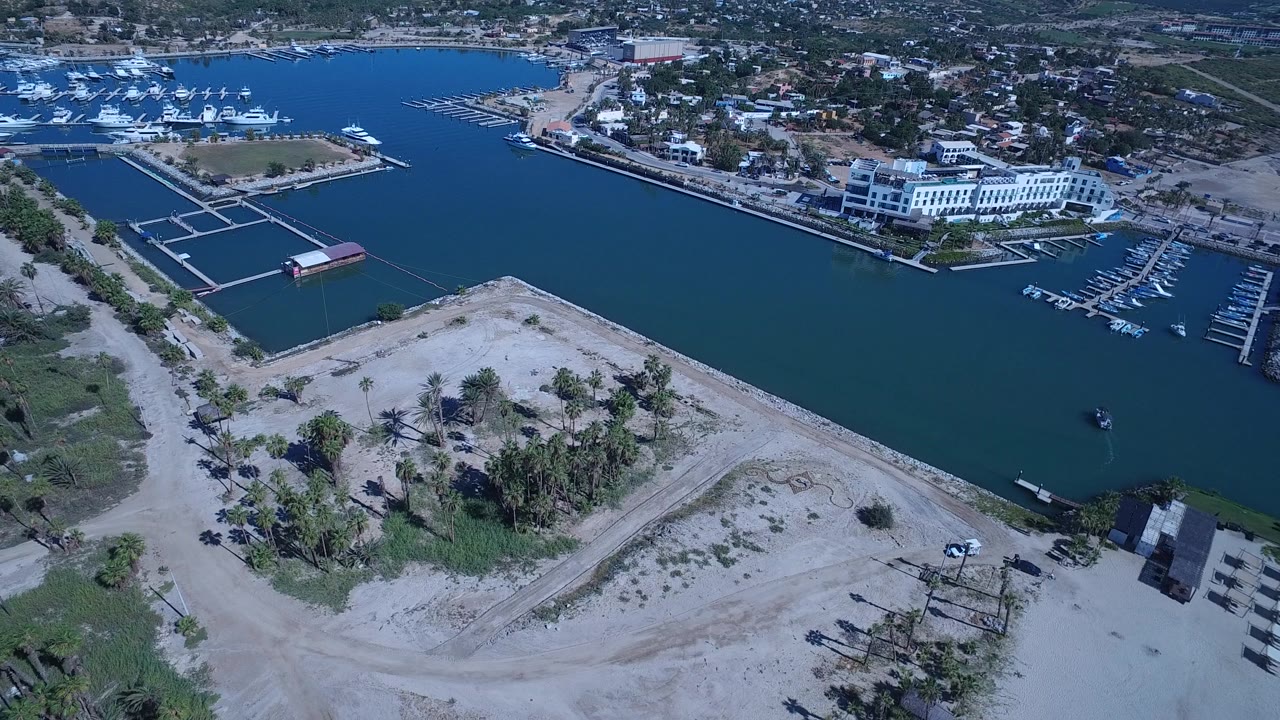 The aerial view in San Jose del Cabo holds over the marina, with a sandy lot in the foreground, rows of boat slips, coastal buildings, and no visible people.