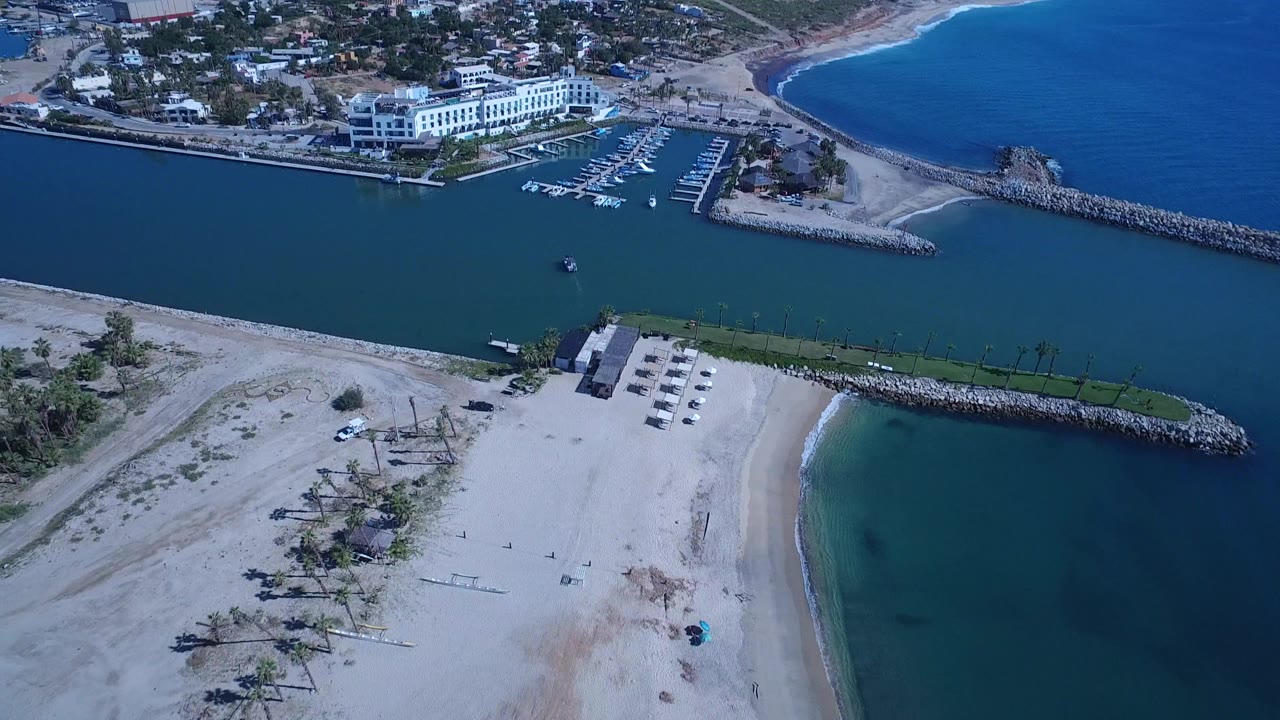 High aerial frame in San Jose del Cabo, Mexico with the beach and inlet filling the lower view, a rock jetty crossing the water, distant buildings, and no visible people.