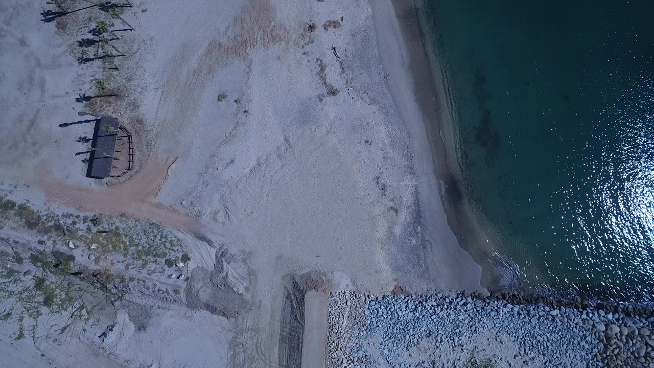 Overhead view of pale beach beside green water and a rocky breakwater.