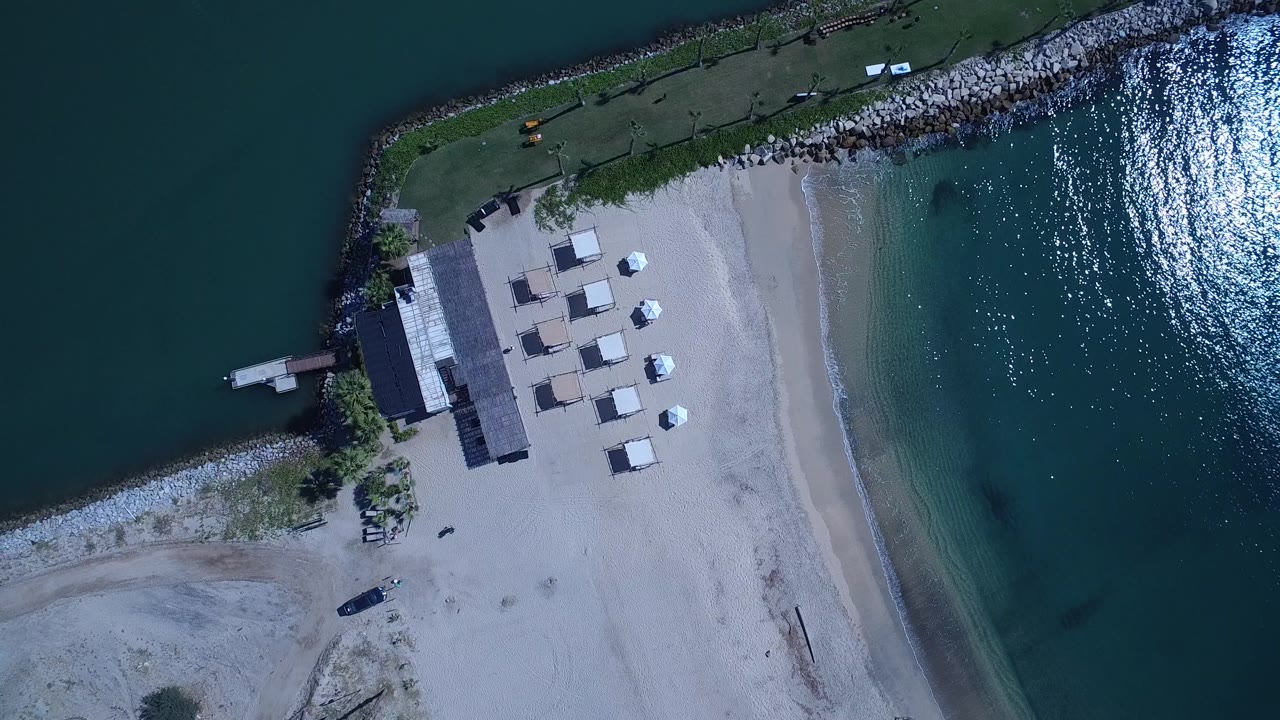 Aerial view over San Jose Del Cabo showing the sandy beach and shade structures shifted slightly within frame, with the rock point and calm blue water around them, no visible people.
