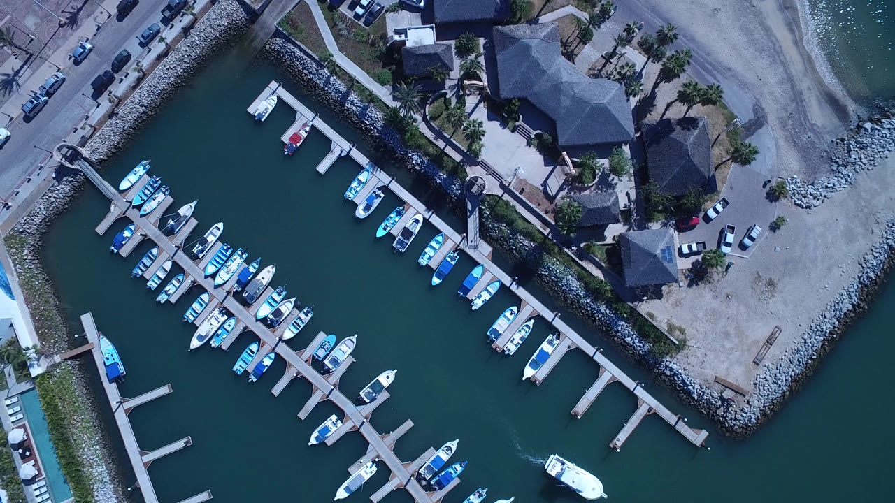 Overhead San Jose del Cabo marina frame showing blue-green water, white boats on both sides of the dock, shoreline rocks, nearby buildings, and no visible people.