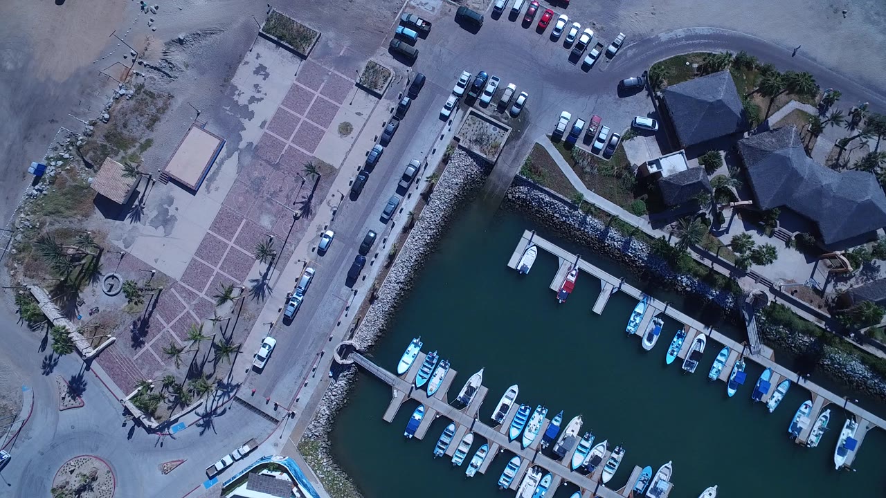 Overhead San Jose del Cabo frame with rows of small boats tied to docks, a rock-lined edge, parked cars along the road, and tiny pedestrians visible near the beach access area.