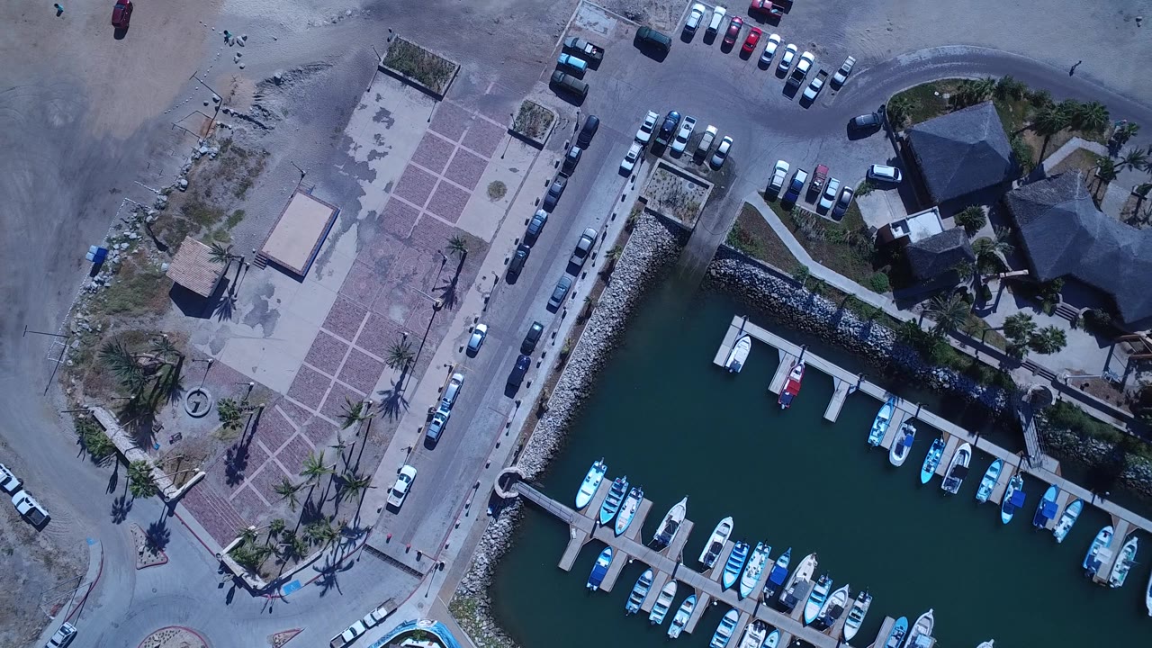 Near-vertical aerial view in San Jose del Cabo with the marina slips on the lower right, parked cars above the waterfront, plaza paving to the left, and tiny people visible on the sand and walkway.