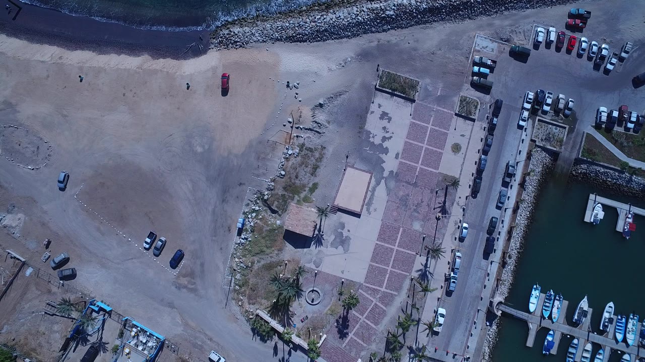Overhead view in San Jose del Cabo, Mexico, showing dark surf at the top, pale beach, parked cars, a paved plaza, and small boats in a marina basin.