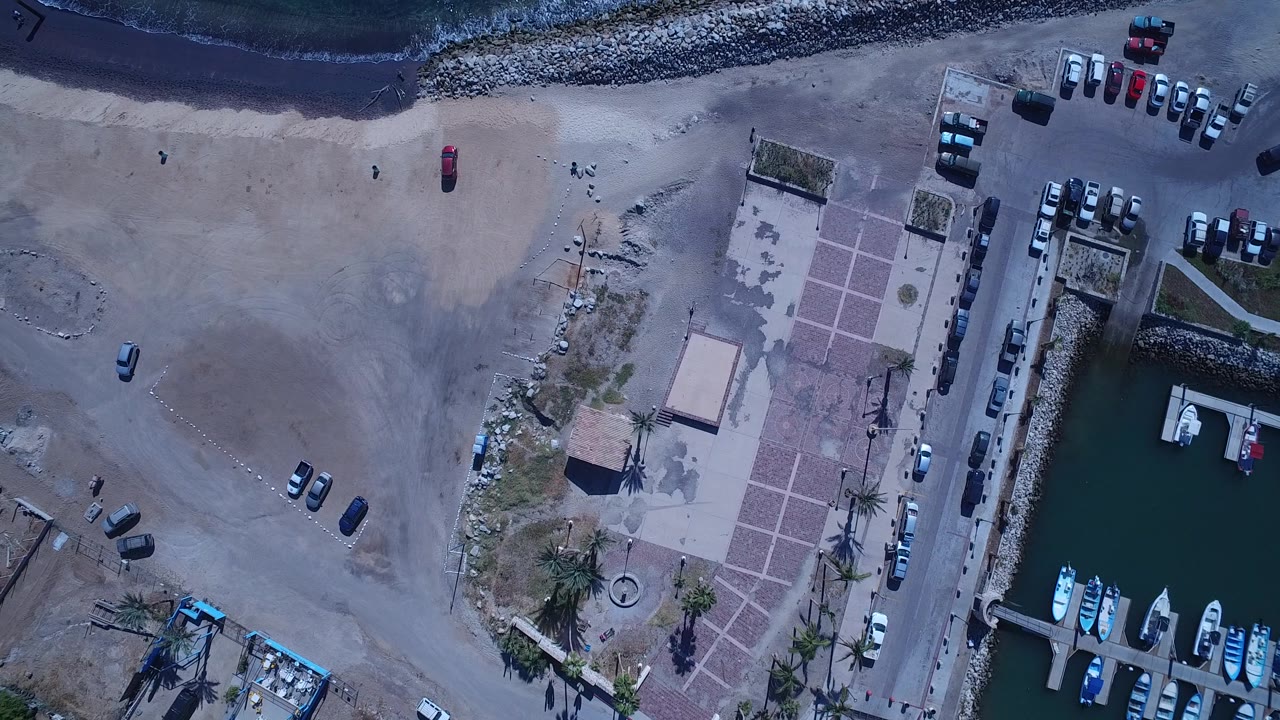 Aerial frame over San Jose del Cabo with the breakwater and surf along the top edge, open sand in the center, cars near the beach, and boats docked at the right.