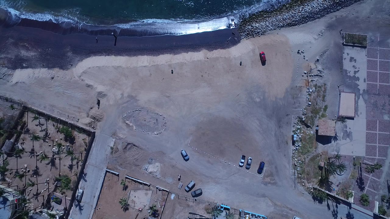 Overhead view in San Jose del Cabo, Mexico, with dark surf along the top, pale sandy lots, parked cars, palm-filled walled properties, and tiny people visible near the shoreline.