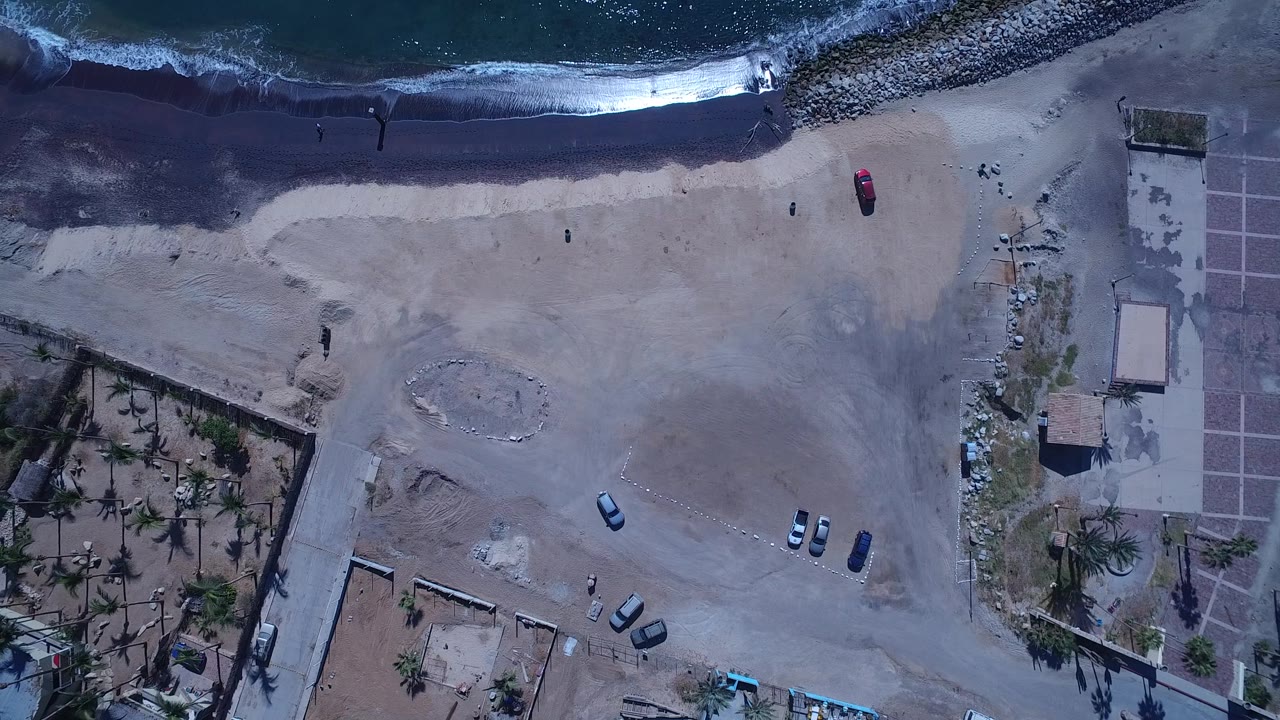 Aerial overhead frame in San Jose del Cabo showing foamy surf, a broad sandy area, several parked cars, a road crossing the lower area, palm trees, and small distant people.