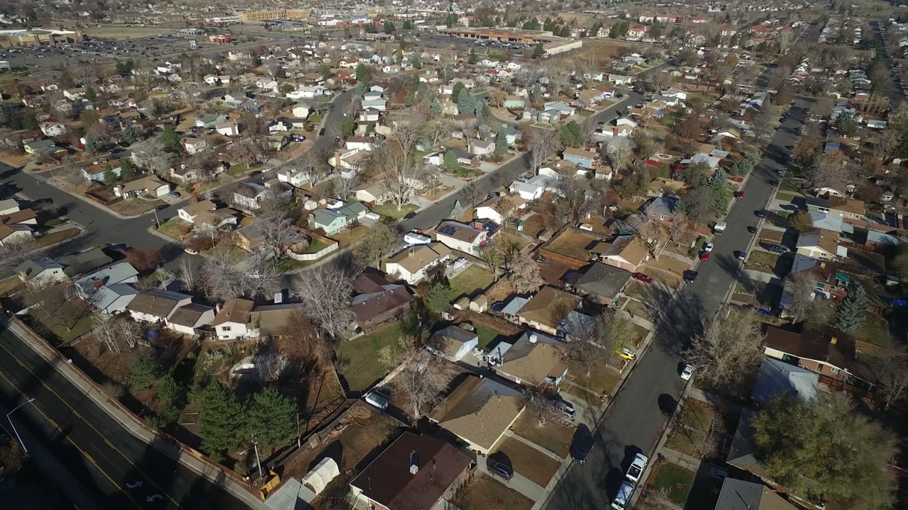 High aerial frame over Westminster, Colorado showing roofs, fenced backyards, bare trees, a main road, and small cars, with no visible people.