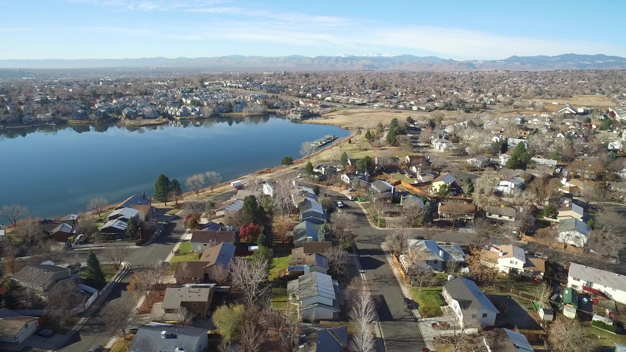 Aerial frame over Westminster, Colorado showing a blue lake on the left, small houses and leafless trees in the foreground, distant neighborhoods and mountains, with no visible people.