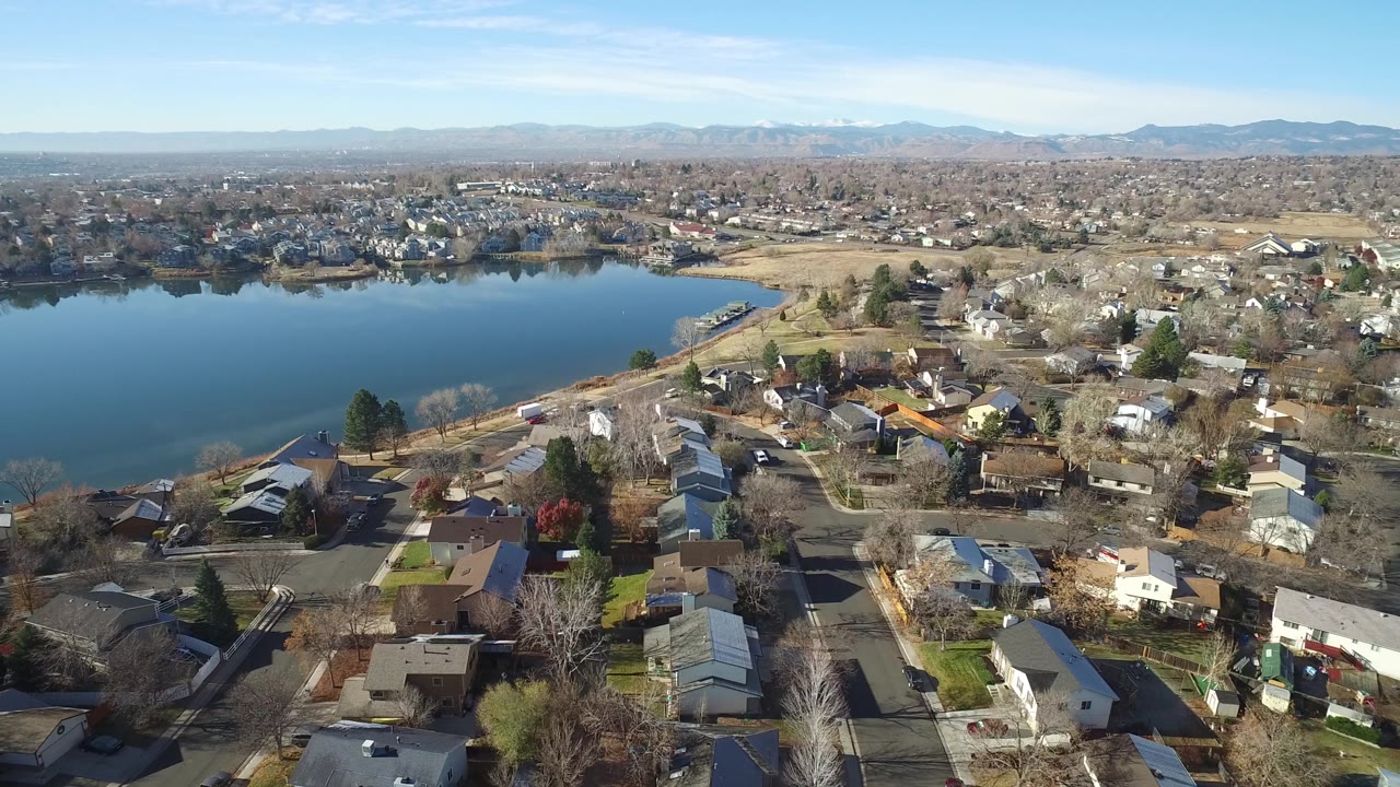 Aerial view in Westminster, Colorado with the lake filling the left side, rooftops and yards below, a broad suburban grid beyond, and no visible people.