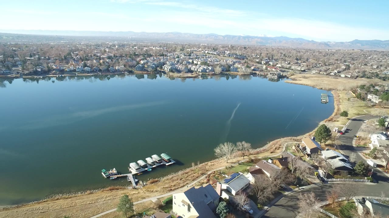 Aerial view in Westminster, Colorado, showing the dock cluster slightly lower in frame, a curved tan shoreline, nearby roofs, broad open water, far-bank houses, and no visible people.