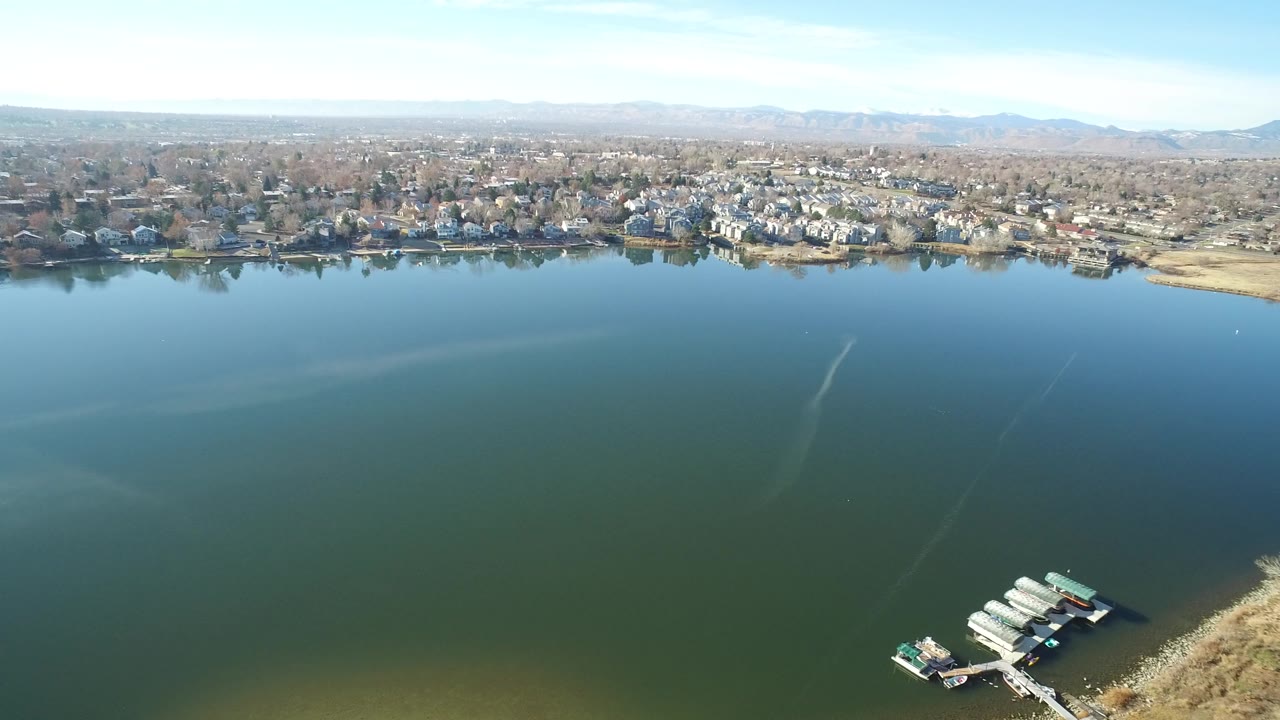 Aerial view over green lake water toward suburban homes and distant mountains.