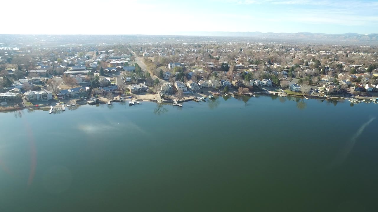 Elevated Westminster, Colorado lake view with small docks along the shore, rows of nearby homes, a bright patch of reflected sun on the water, and no visible people.