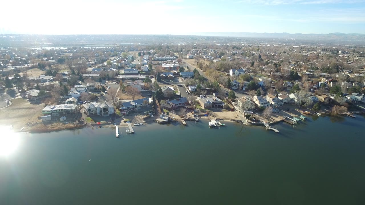Wide aerial view in Westminster, Colorado showing dark lake water, private docks, shoreline houses, residential streets, distant haze, and no visible people.