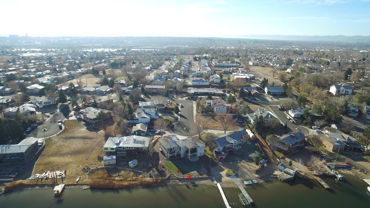 Aerial frame over Westminster, Colorado showing waterfront homes, docks, brown lawns, curving streets, distant suburbs, and no visible people.