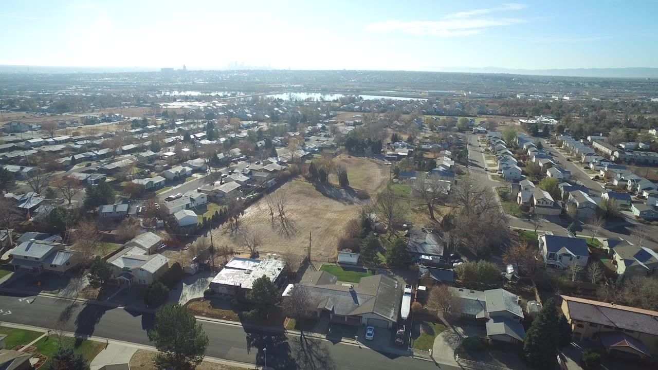 High aerial view over Westminster, Colorado houses, leafless trees, pale open fields, distant water, and a hazy skyline, with no visible people.