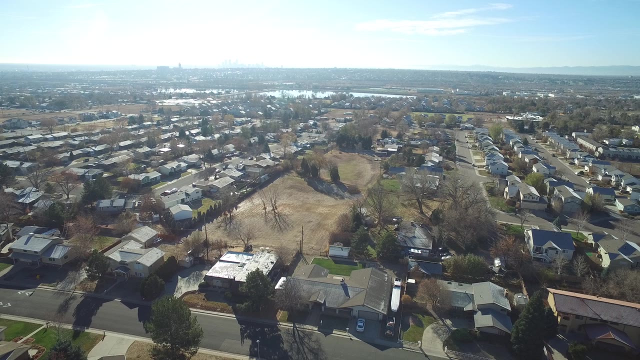 Aerial frame from Westminster, Colorado showing rooflines, backyard lots, open brown grass, distant water, and a low urban horizon, with no visible people.