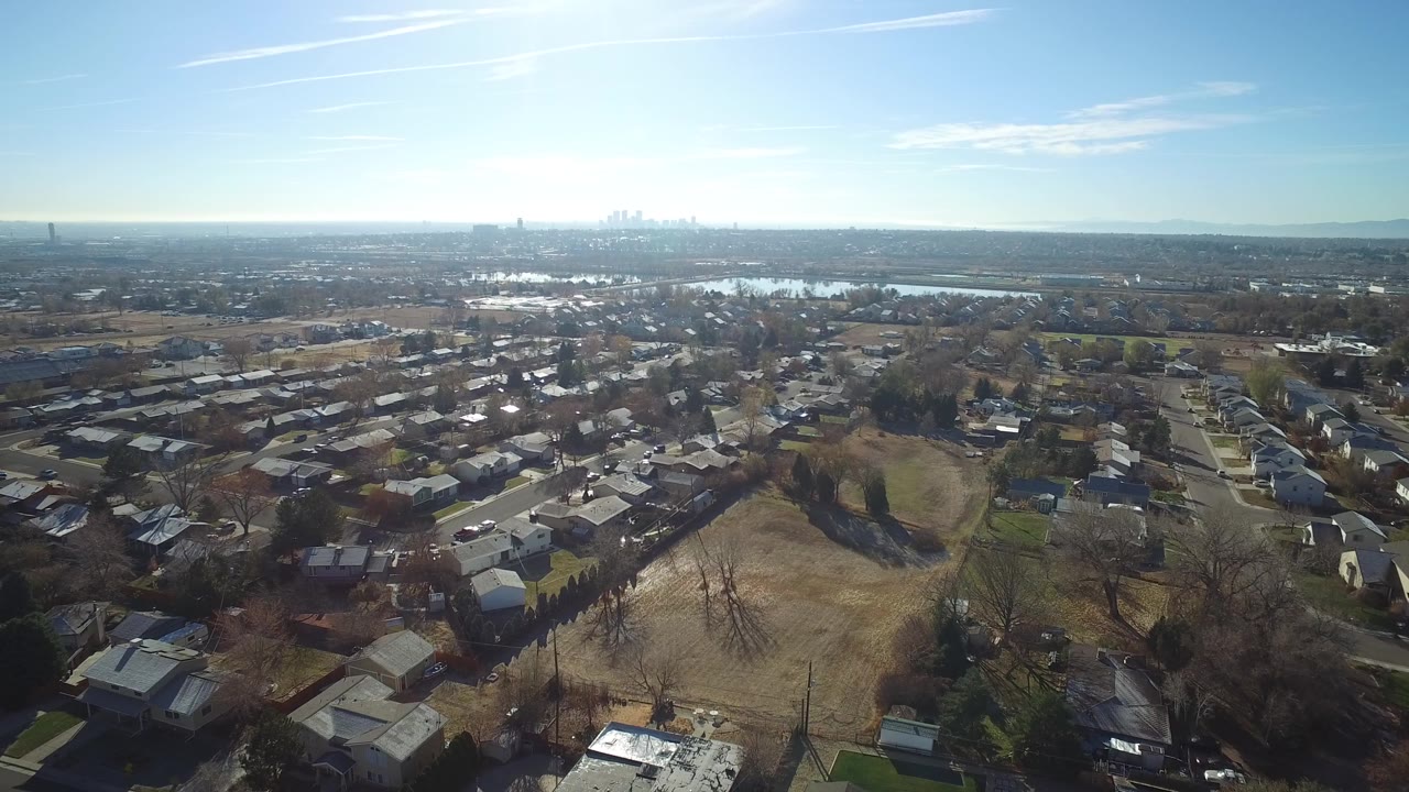 A wide Westminster, Colorado aerial frame shows residential rooftops below, an open tan field near the center, water beyond the homes, a bright blue sky, and no visible people.