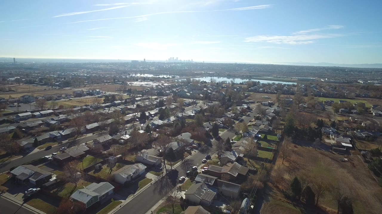 Opening aerial frame over Westminster, Colorado homes and streets, with bare trees, brown open ground, distant water, a hazy skyline, blue sky, and no visible people.