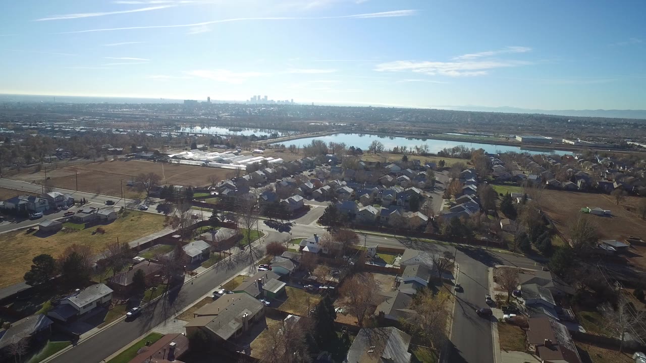 Wide aerial view over Westminster, Colorado suburbs with residential streets in the foreground, reflective water beyond the homes, a distant skyline, and no visible people.