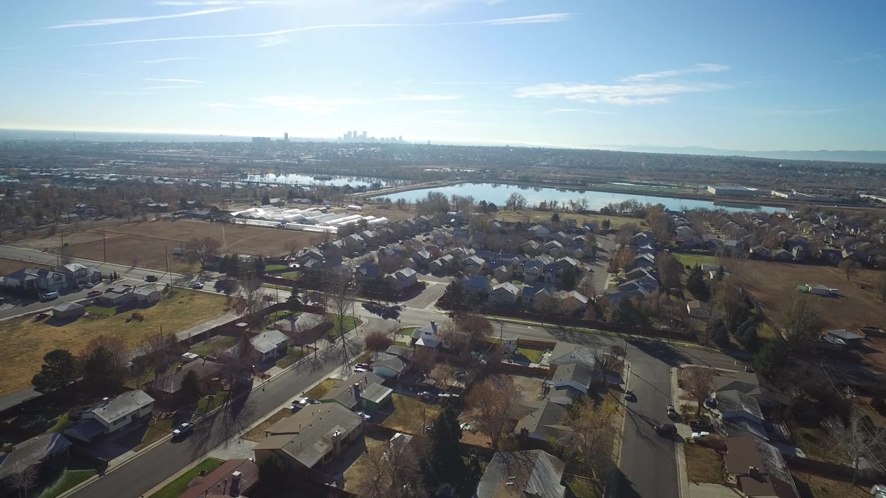 Aerial frame over Westminster, Colorado showing clustered homes, curving streets, open brown fields, a water body, distant buildings, and no visible people.