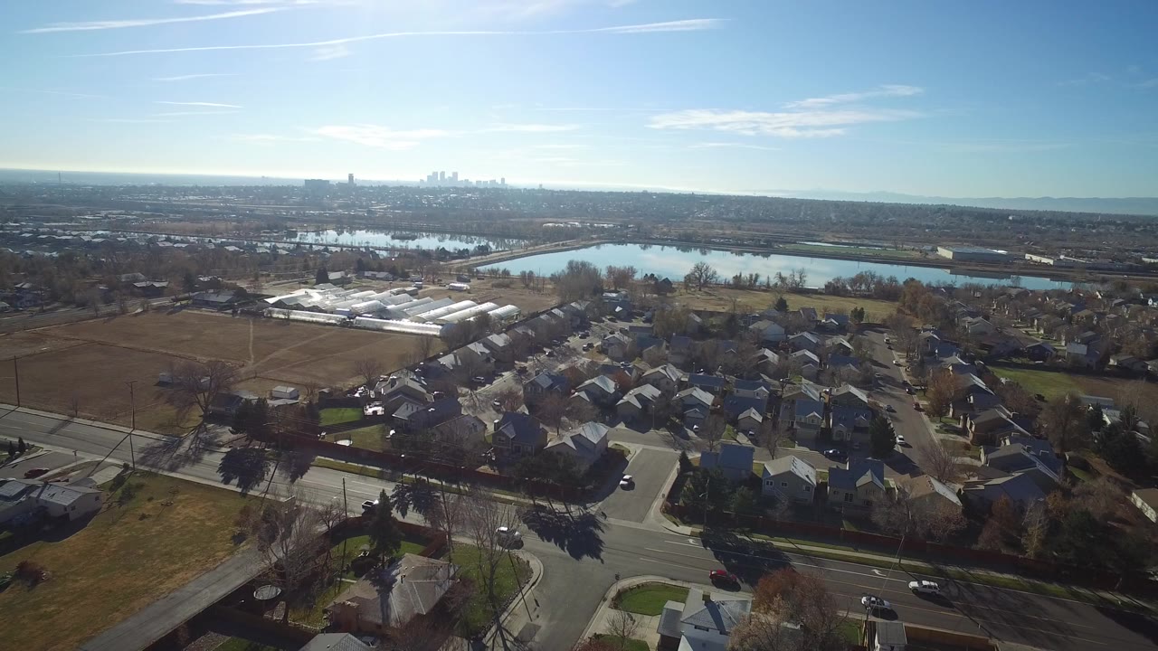 Aerial view of suburban houses, roads, fields, trees, water, and distant skyline.