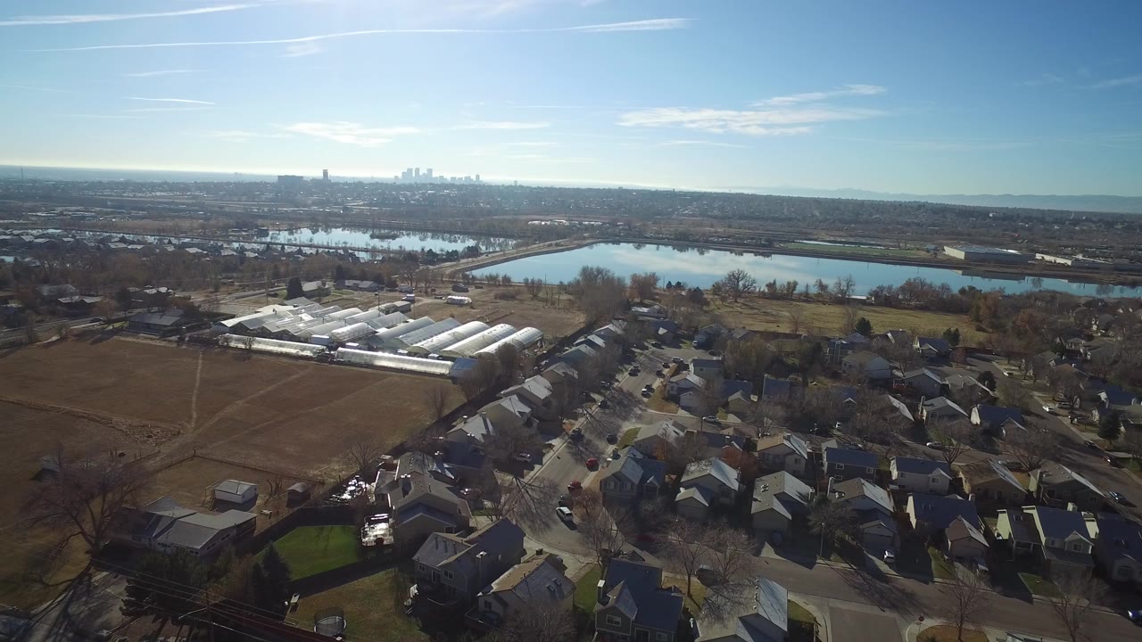 Aerial view over houses, fields, lakes, and distant skyline under a blue sky.