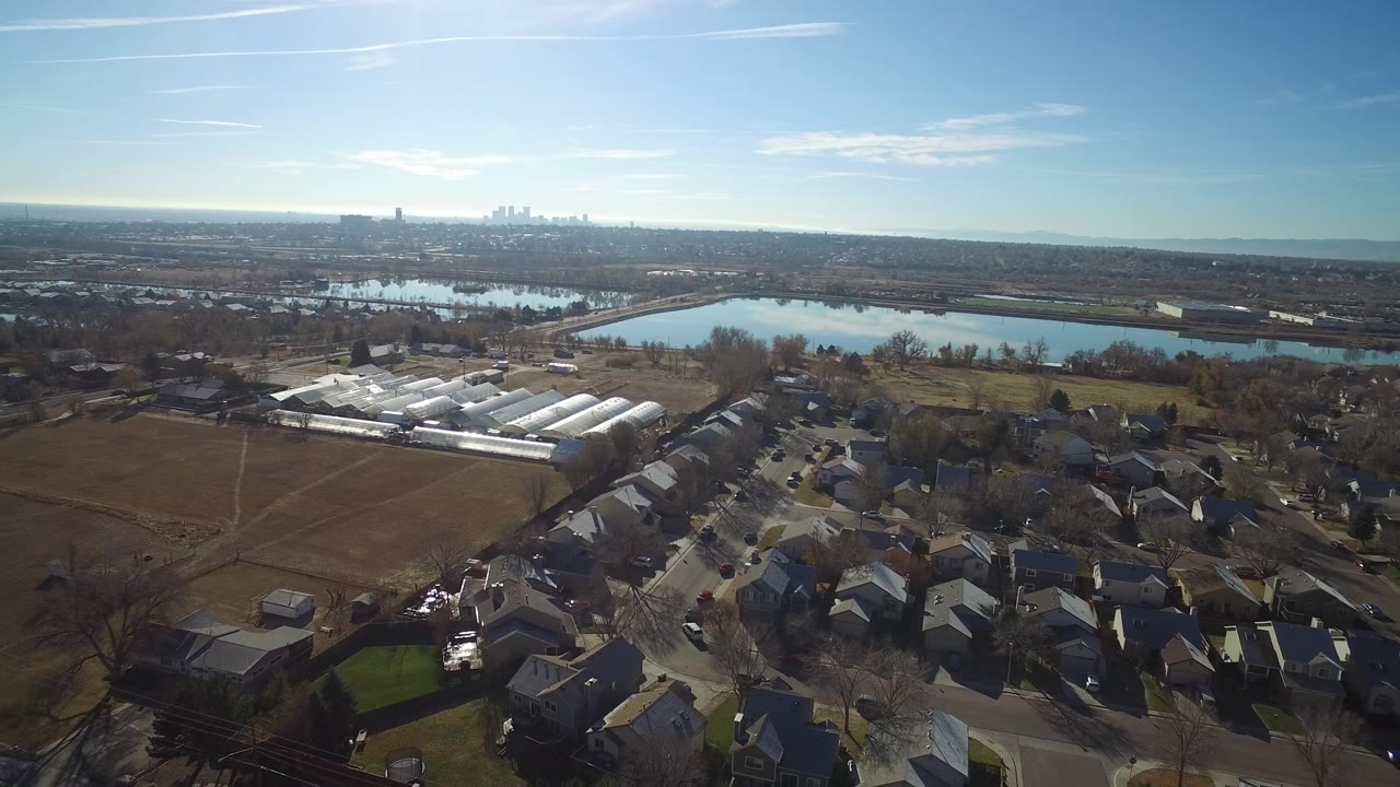 Second frame over Westminster, Colorado with the residential street pattern lower in frame, white greenhouse-like roofs left of center, open fields, lakes across the middle distance, far skyline, and no visible people.