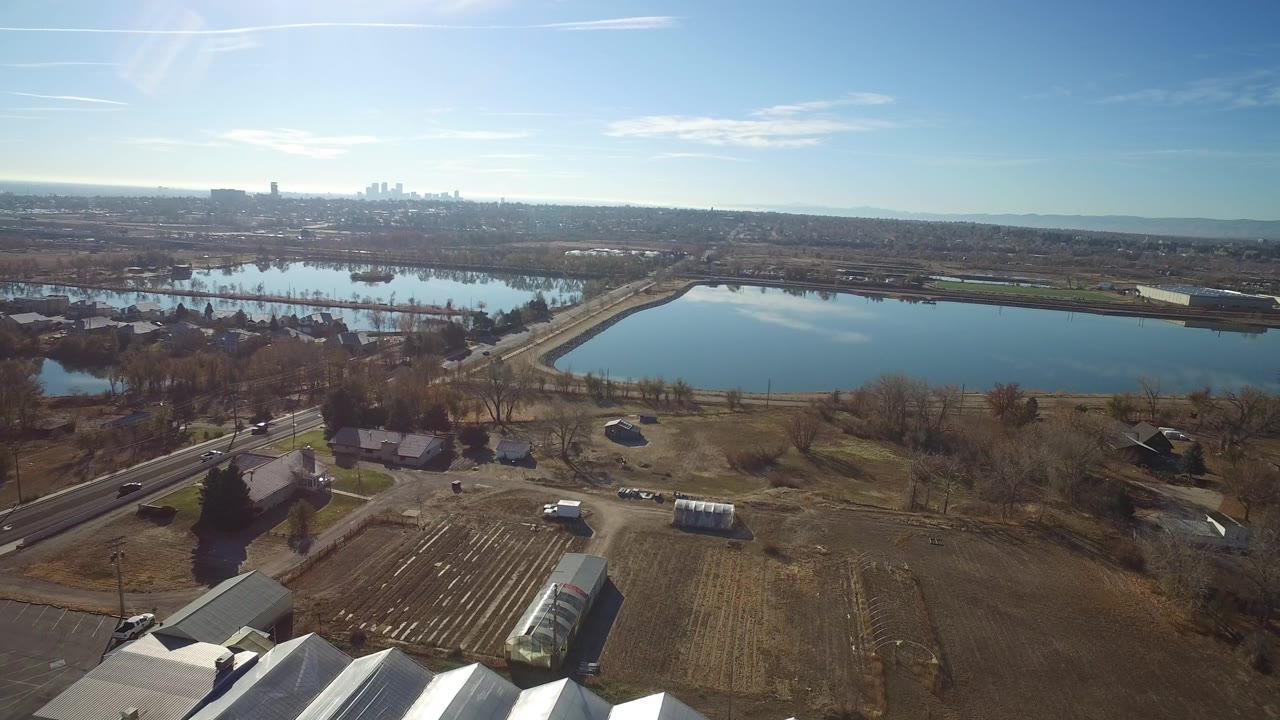The aerial view in Westminster, Colorado looks across foreground farm-like plots and roofs toward a reflective lake, roads, bare trees, and far urban towers with no visible people.