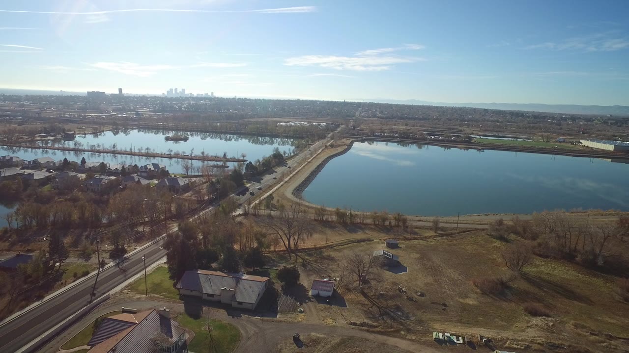 A high aerial frame over Westminster, Colorado shows a blue lake, curving road, scattered homes, bare trees, and a distant skyline under bright daylight, with no visible people.
