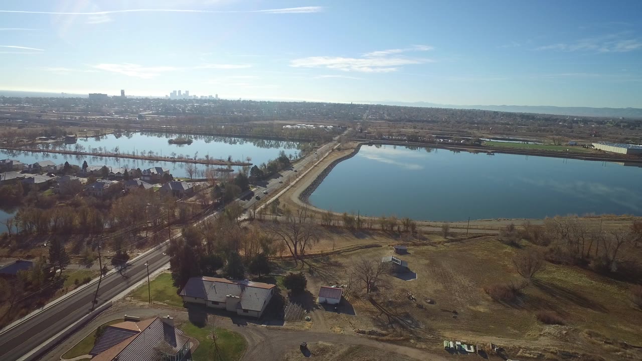 The next frame keeps the Westminster lake centered while the road, clustered homes, leafless trees, and far skyline sit under strong sun haze, with no visible people.