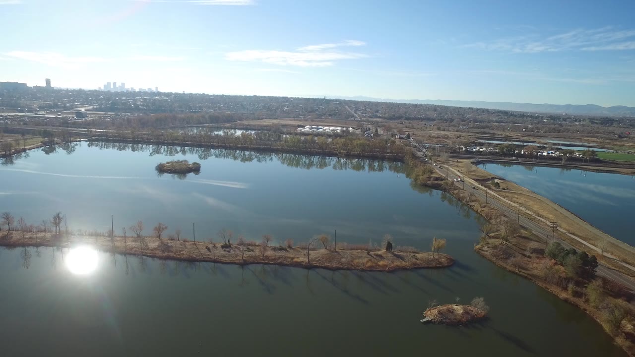 Wide drone view in Westminster, Colorado with calm water on both sides of a dividing road, a small island left of center, shoreline vegetation, and no visible people.