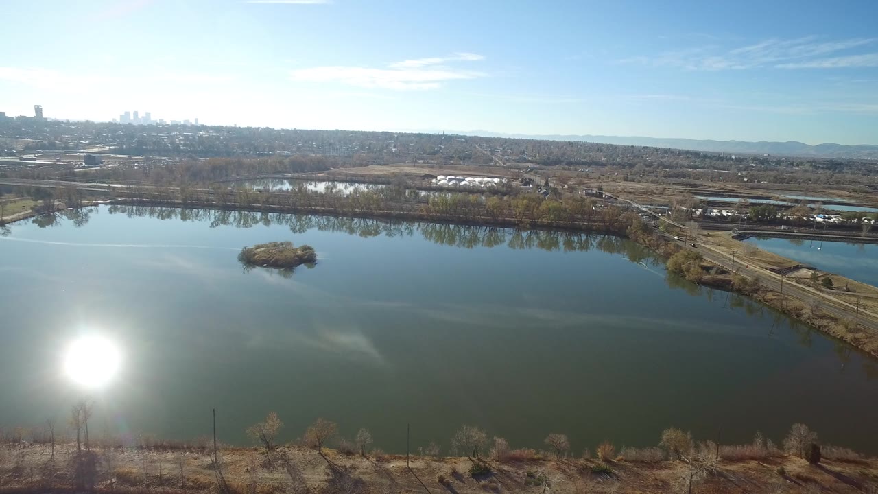 Aerial view of a lake with sun glare, a small island, shoreline trees, and distant buildings.