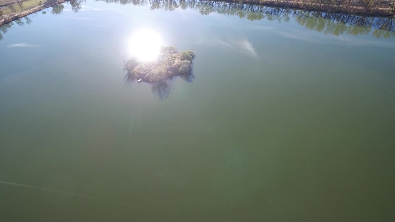 Drone view of a brushy island beside a large sun reflection on a lake.