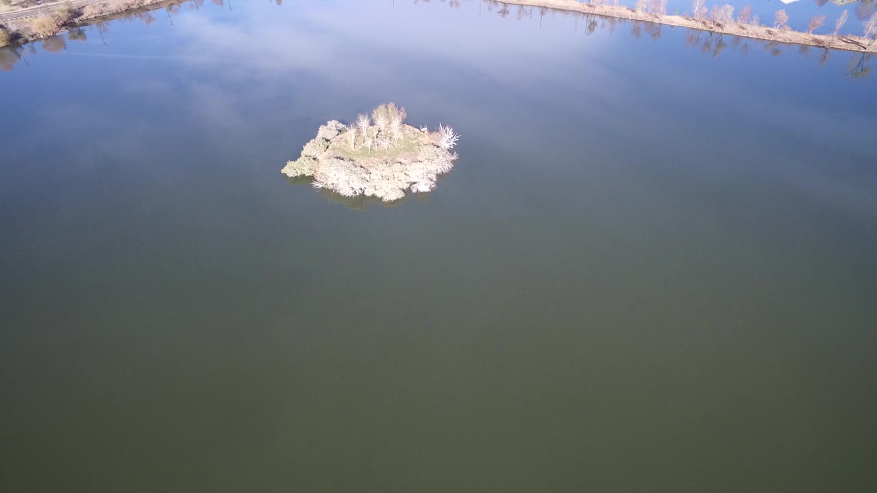 Westminster, Colorado aerial view with the small island slightly right of center, smooth water below it, pale cloud reflection above, and no visible people.