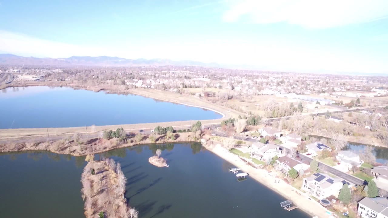 Aerial frame in Westminster, Colorado, with a large lake at left, a darker foreground pond, clustered homes, leafless trees, and hazy mountains in the distance, no visible people.