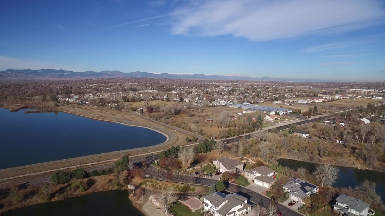 A wide aerial frame over Westminster, Colorado shows a curved artificial lake at left, roads and houses below, distant foothills on the horizon, and no visible people.