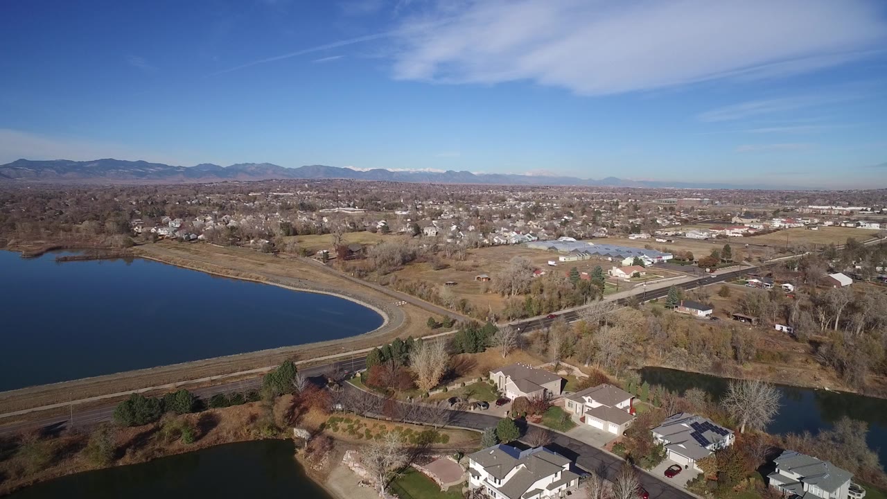 An overhead view in Westminster, Colorado looks across the lake shoreline toward a road corridor, scattered homes, open ground, industrial-looking buildings, and no visible people.